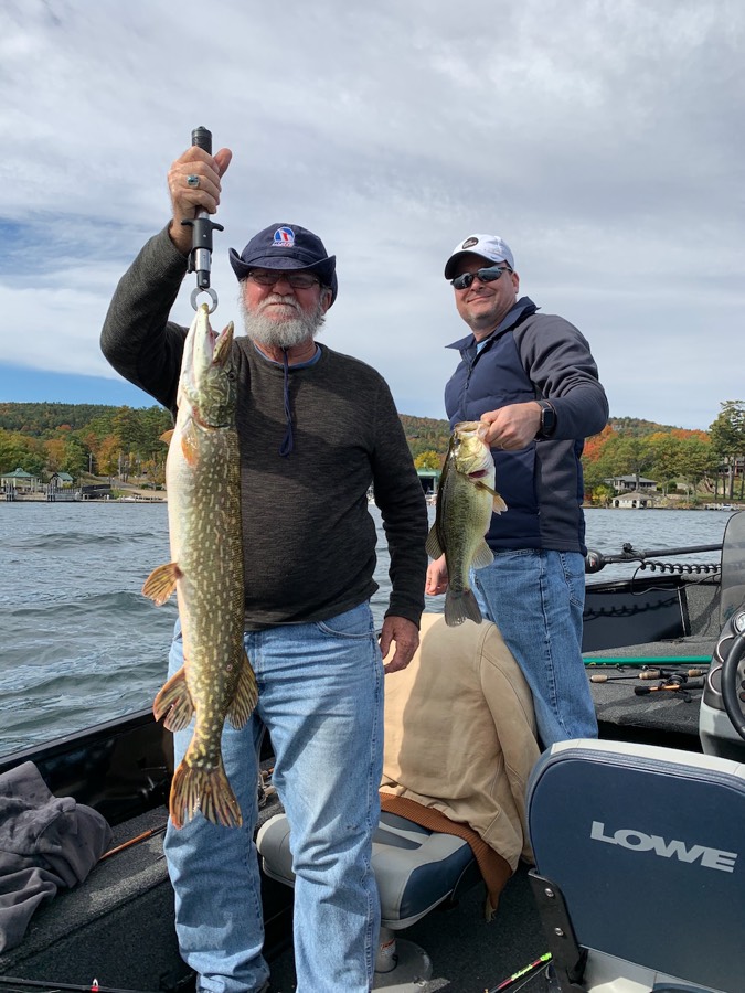 Donald Barde holding a northern pike and Brian Barde holding a largemouth bass, standing in a boat on a lake with autumn foliage behind them