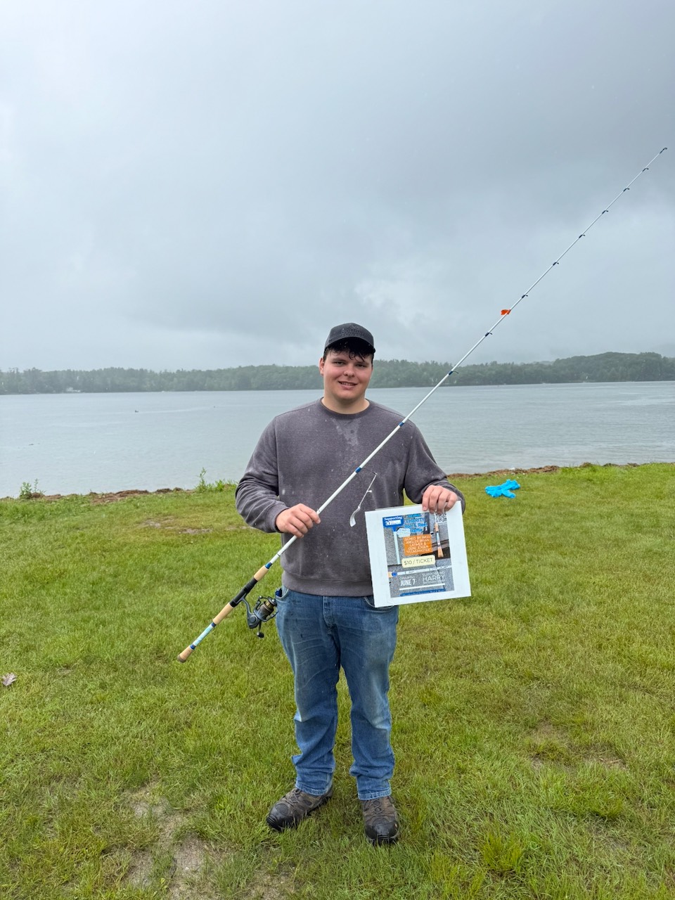 The 2025 Jimmy Fund Rod winner holding his rod and a raffle flyer on an overcast day by the water